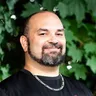 Joshua Miller smiling outdoors in front of green leaves, wearing a black LezFindr t-shirt